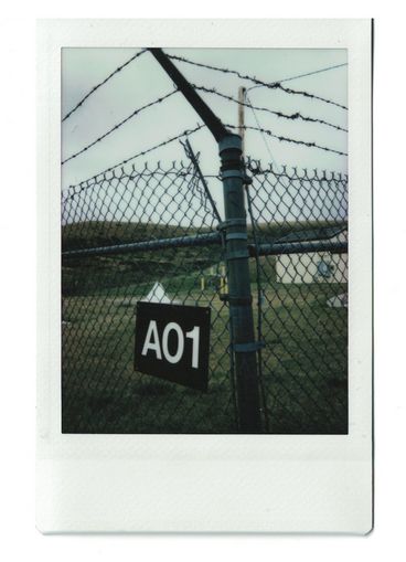 Polaroid eines Maschendrahtzauns mit einem Schild „A01“, dahinter grüne Felder und ein Haus.