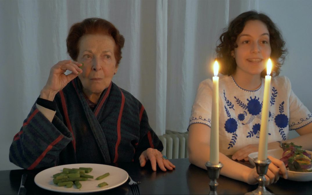 [Translate to English:] Filmstill aus FASHIONS: Eine junge und eine ältere Frau sitzen nebeneinander an einem Esstisch, vor sich ihre Teller mit Essen. Auf dem Tisch stehen zwei Kerzen.