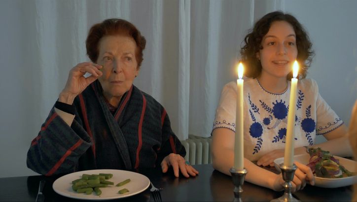[Translate to English:] Filmstill aus FASHIONS: Eine junge und eine ältere Frau sitzen nebeneinander an einem Esstisch, vor sich ihre Teller mit Essen. Auf dem Tisch stehen zwei Kerzen.