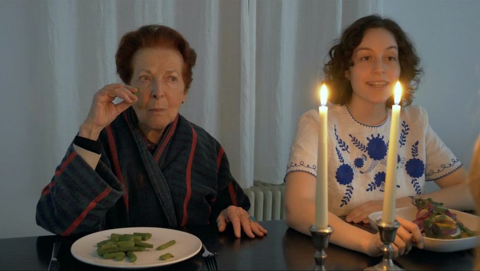 [Translate to English:] Filmstill aus FASHIONS: Eine junge und eine ältere Frau sitzen nebeneinander an einem Esstisch, vor sich ihre Teller mit Essen. Auf dem Tisch stehen zwei Kerzen.