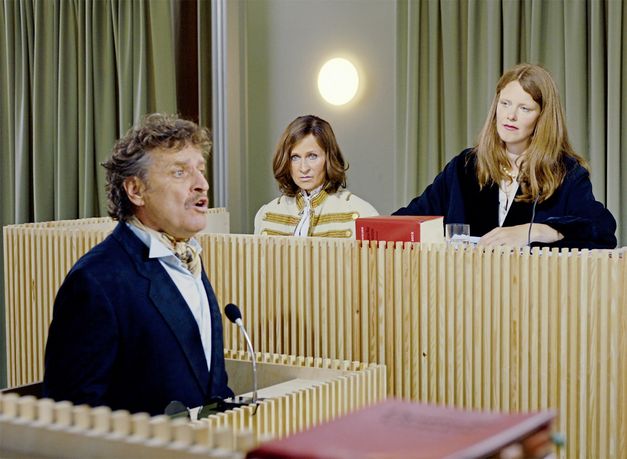 Still from the film "L’état et moi" by Max Linz. A court scene: A man sits in the witness stand and speaks. In the background we see a female lawyer, next to her a woman in an old-fashioned uniform jacket.