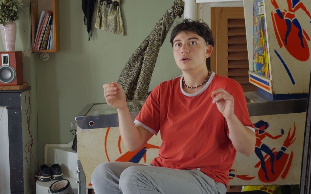 A person sits in a room raising both hands, surrounded by furniture, fabrics, and patterned objects.