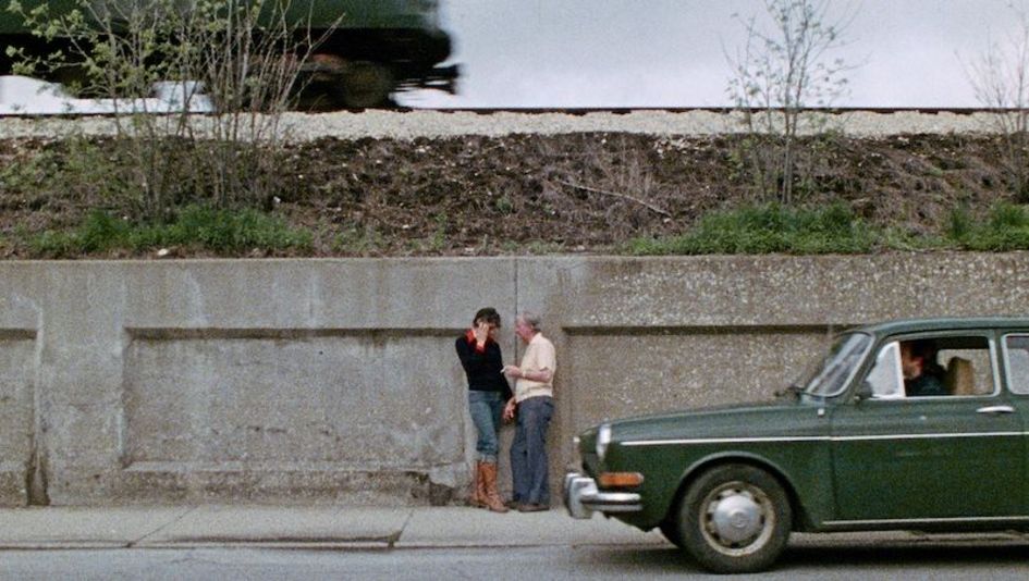 Filmstill aus ELEVEN BY FOURTEEN: Zwei Personen stehen an einer Mauer und unterhalten sich. Vor ihnen fährt ein grünes Auto über die Straße.