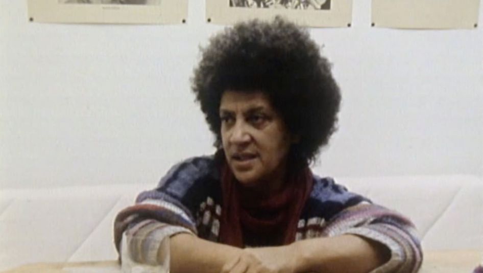 Film still from FASIA JANSEN – SÄNGERIN: Fasia Jansen is sitting at a table. There is a glass of water in front of her, and her arms are resting on the table.