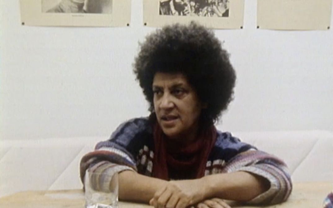 Film still from FASIA JANSEN – SÄNGERIN: Fasia Jansen is sitting at a table. There is a glass of water in front of her, and her arms are resting on the table.