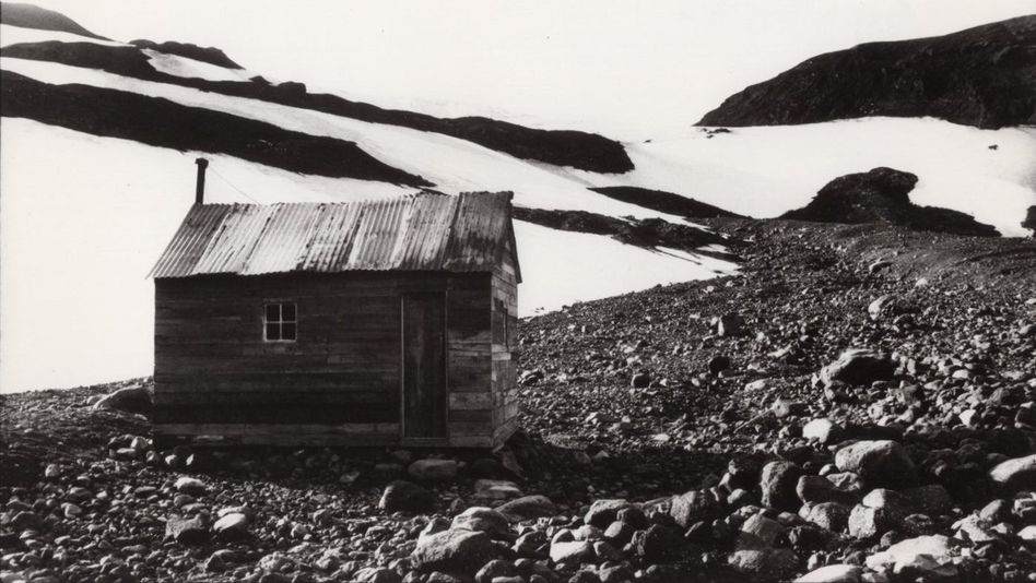 Filmstill aus THE GOLD DIGGERS: Eine kleine Hütte aus Brettern und einem Wellblechdach steht auf einem bergigen Geröllfeld. Im Hintergrund ist Schnee zu sehen.