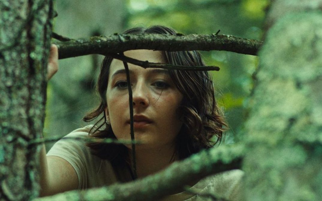 Film still from GOOD ONE: A teenage girl peers out from behind the branches of a tree.