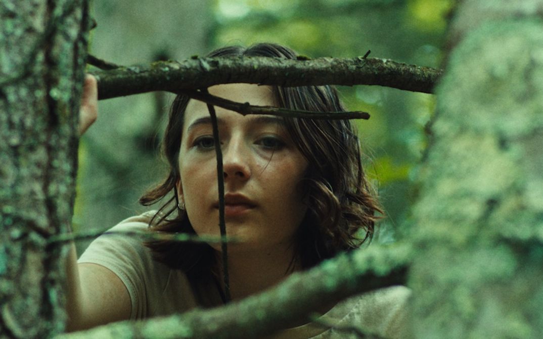 Film still from GOOD ONE: A teenage girl peers out from behind the branches of a tree.