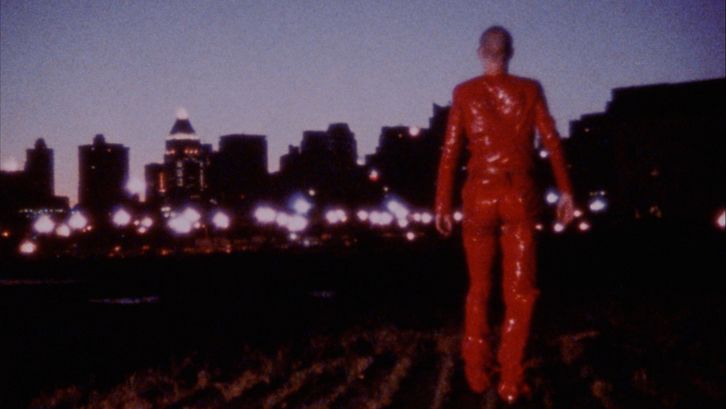 Filmstill aus ROTE OHREN FETZEN DURCH ASCHE: Eine ganz in Rot gekleidete Person geht in der Dunkelheit auf die Lichter einer Stadt zu.