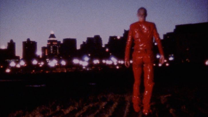 Film still from ROTE OHREN FETZEN DURCH ASCHE: A person dressed entirely in red walks towards the lights of a city in the dark.