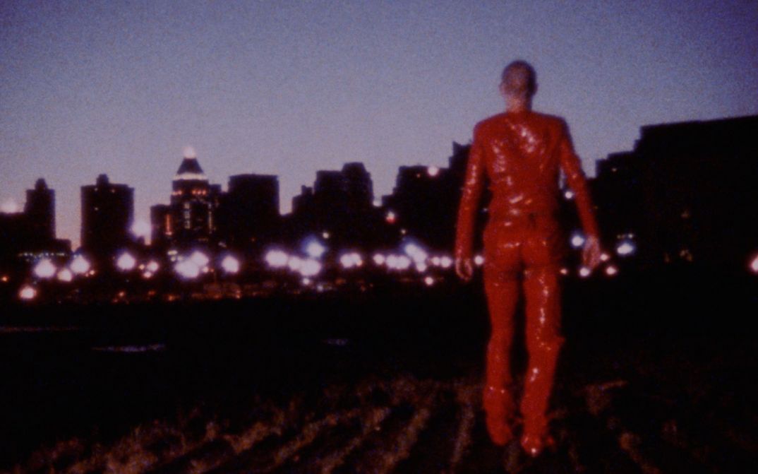 Filmstill aus ROTE OHREN FETZEN DURCH ASCHE: Eine ganz in Rot gekleidete Person geht in der Dunkelheit auf die Lichter einer Stadt zu.