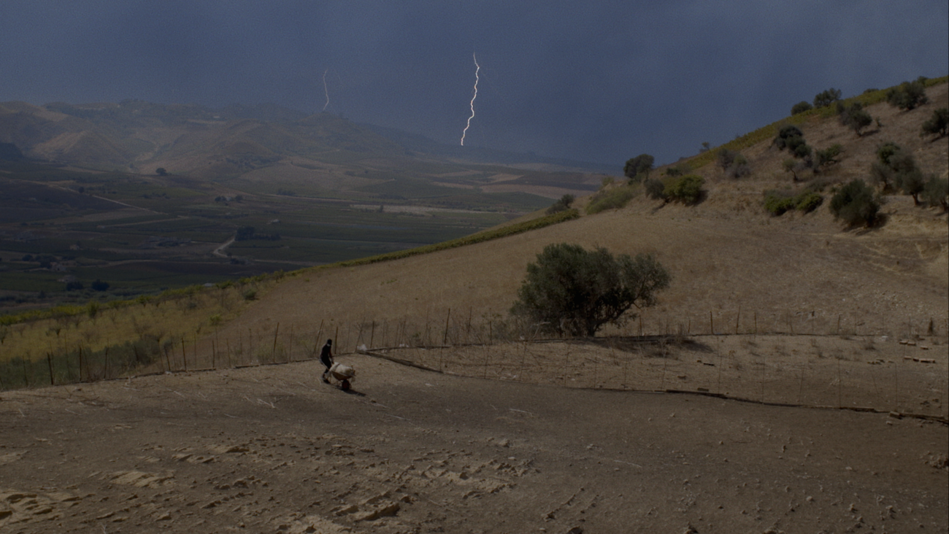 Film still from TARDO AGOSTO: A man is working in a field in a hilly landscape, a flash of lightning can be seen in the sky.