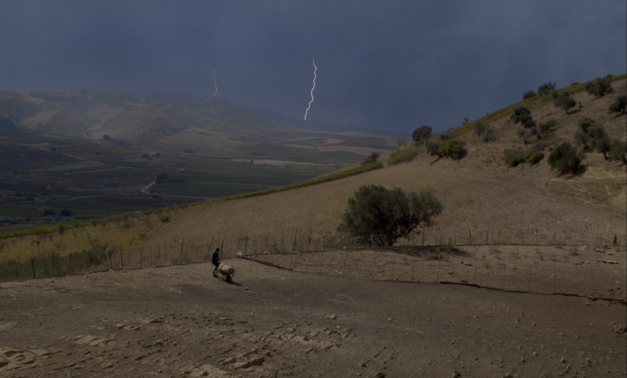 Filmstill aus TARDO AGOSTO: Ein Mann arbeitet auf einem Feld in einer hügeligen Landschaft, im Himmel ist ein Blitz zu sehen.