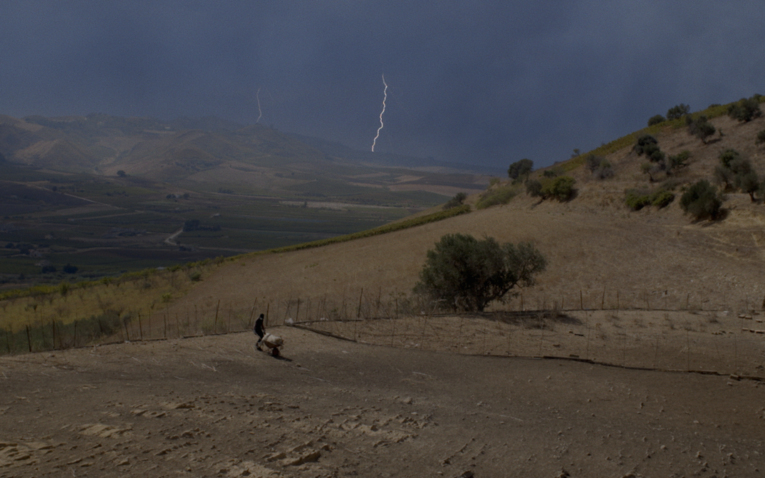 Film still from TARDO AGOSTO: A man is working in a field in a hilly landscape, a flash of lightning can be seen in the sky.