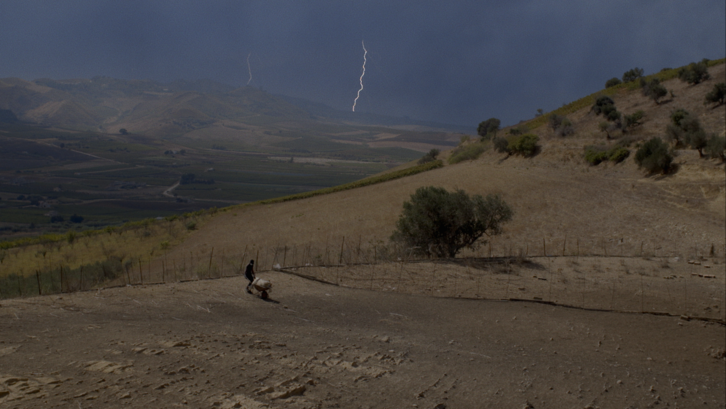 Film still from TARDO AGOSTO: A man is working in a field in a hilly landscape, a flash of lightning can be seen in the sky.