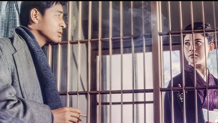 Filmstill aus AKITSU SPRINGS: Ein Mann und eine Frau blicken sich durch ein Gitter hindurch an.