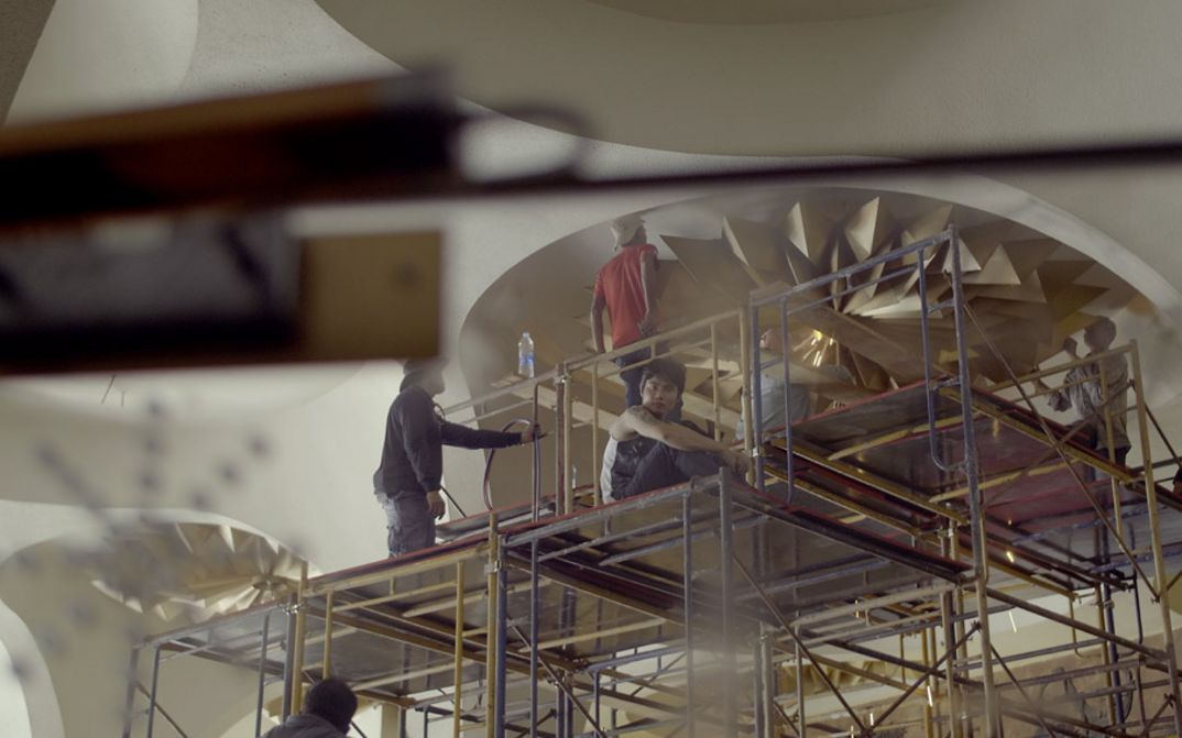 Still from the film "Scala" by Ananta Thitanat. We see a shot from below as a group of men on scaffolding work to remove light fixtures from the ceiling.