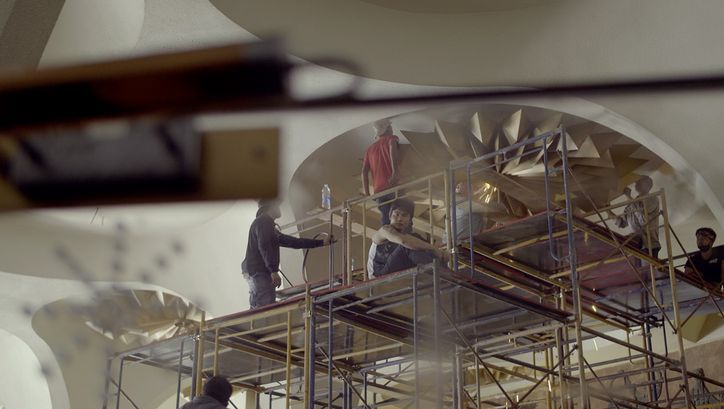 Still from the film "Scala" by Ananta Thitanat. We see a shot from below as a group of men on scaffolding work to remove light fixtures from the ceiling.