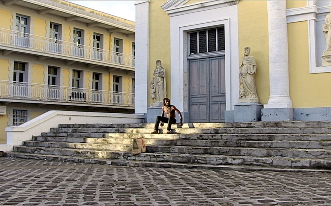 Filmstill aus "L’ homme-vertige" von Malaury Eloi Paisley. Zu sehen ist ein oberkörperfreier Mann auf einer Treppe vor einem Gebäude. Das Gebäude hat eine gelbe Fassade. Links und rechts vom Eingang stehen Statuen.