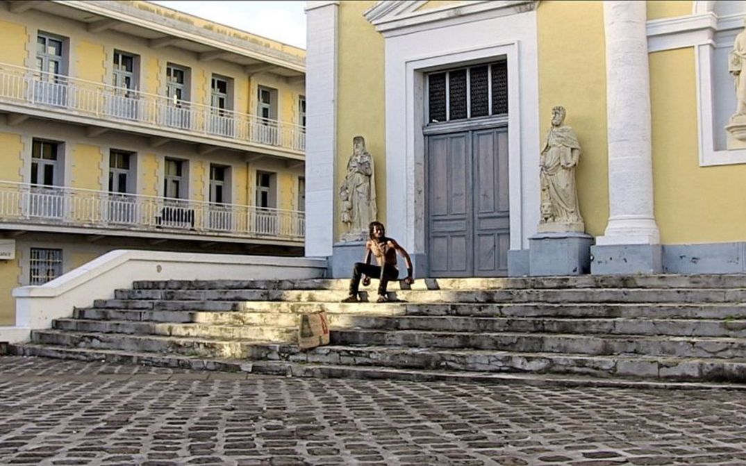 Filmstill aus "L’ homme-vertige" von Malaury Eloi Paisley. Zu sehen ist ein oberkörperfreier Mann auf einer Treppe vor einem Gebäude. Das Gebäude hat eine gelbe Fassade. Links und rechts vom Eingang stehen Statuen. 
