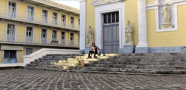 Film still from "L’ homme-vertige" by Malaury Eloi Paisley. It shows a shirtless man on a staircase in front of a building. The building has a yellow façade. There are statues to the left and right of the entrance.
