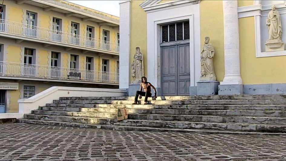 Film still from "L’ homme-vertige" by Malaury Eloi Paisley. It shows a shirtless man on a staircase in front of a building. The building has a yellow façade. There are statues to the left and right of the entrance.