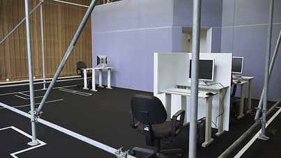 Still from the film "77sqm_9:26min" by Forensic Architecture. A room with metal scaffolding and three computer work stations. 