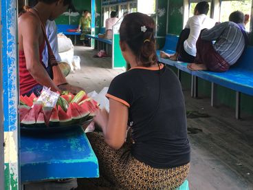 A behind-the-scenes shot of a few people sitting on blue benches. A bowl of watermelon slices is placed on the left bench.