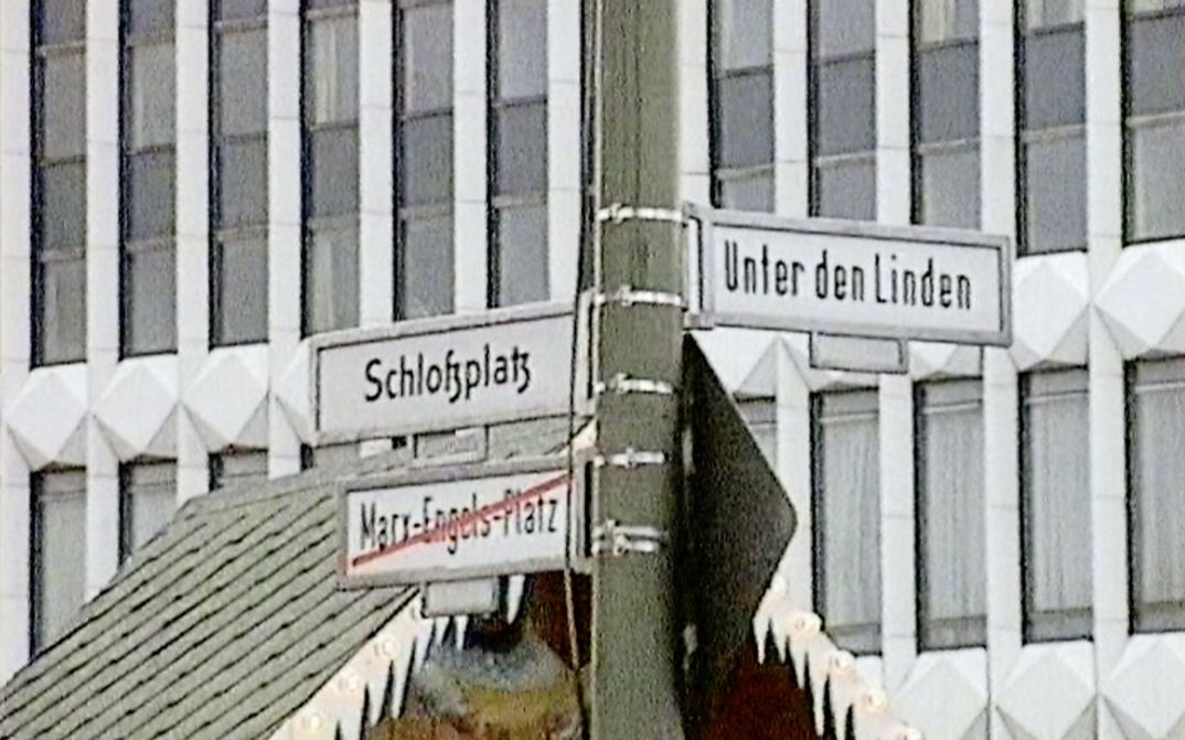 [Translate to English:] Filmstill aus „Die leere Mitte“ von Hito Steyerl. Straßenschilder in Berlin: Schlossplatz, Unter den Linden und durchgestrichen Marx-Engels-Platz. 