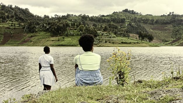 Stillfilm aus „The Bride" von Myriam U. Birara. Zwei Frauen mit dem Rücken zur Kamera sind am Ufer eines Flusses. Sie blicken auf das andere, grüne Ufer.