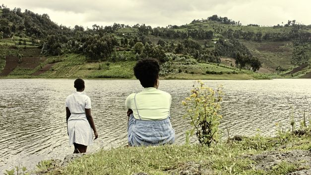 Stillfilm from "The Bride" von Myriam U. Birara. Two women with their backs to the camera are on the bank of a river. They are looking at the other, green bank.