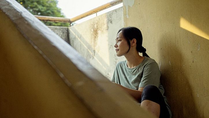 Film still from "Oasis of Now" by Chee Sum Chia. You can see a woman outside in the shade on a staircase in sunny weather.