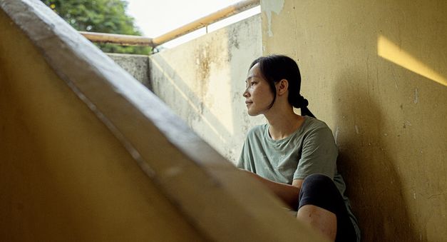 Film still from "Oasis of Now" by Chee Sum Chia. You can see a woman outside in the shade on a staircase in sunny weather.