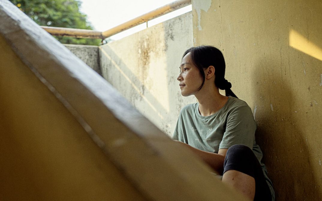 Filmstill aus "Oasis of Now" von Chee Sum Chia. Zu sehen ist eine Frau draußen im Schatten auf einer Treppe bei sonnigem Wetter.