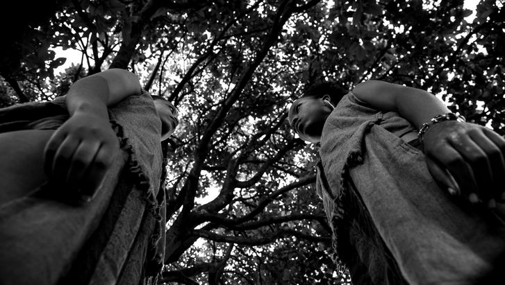 Film still from MAHERE: Black and white image of two people facing each other, taken from an extreme bottom view. Above them can be seen treetops.