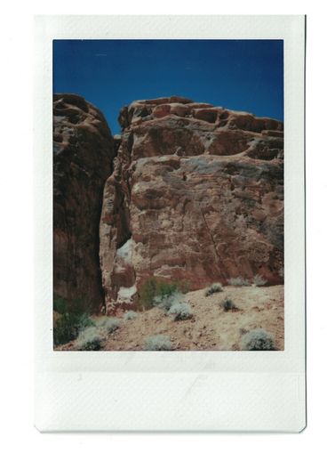 Polaroid of a desert cliff seen from below, with shrubs in the foreground.