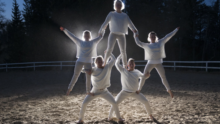 Film still from STUDIES ON THE ECOLOGY OF DRAMA: Five people, all dressed in white, form a small human pyramid. They stand in the dark on a sandy area and are illuminated by a spotlight.
