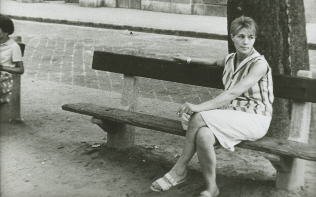 Filmstill aus ENCOUNTER: Eine junge Frau in sommerlichen Kleidung sitzt auf einer Bank und blickt sehnsüchtig in die Ferne.