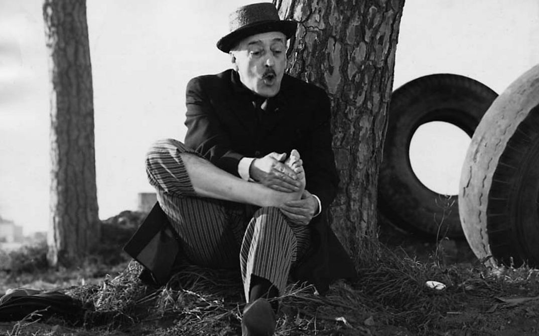 Film still from UCCELLACCI E UCCELLINI. A man sits on the street holding his aching foot.