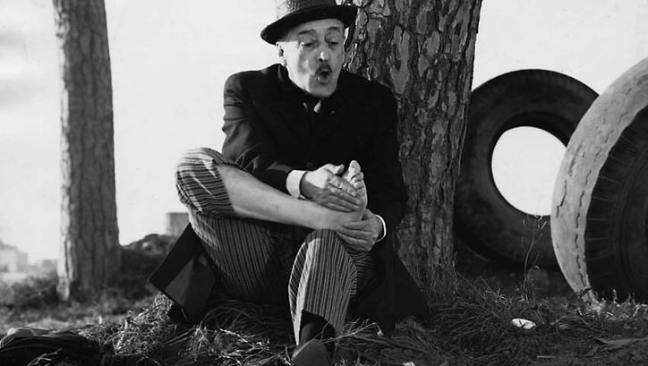 Filmstill aus UCCELLACCI E UCCELLINI. Ein Mann sitzt auf der Straße und hält seinen schmerzenden Fuß in der Hand.