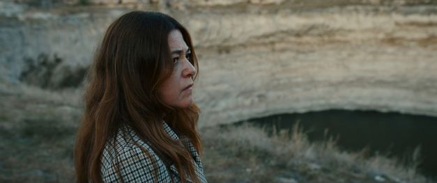 Frau steht im Profil in einer kargen Landschaft und blickt auf eine abgesenkte Fläche oder einen Wasserlauf.