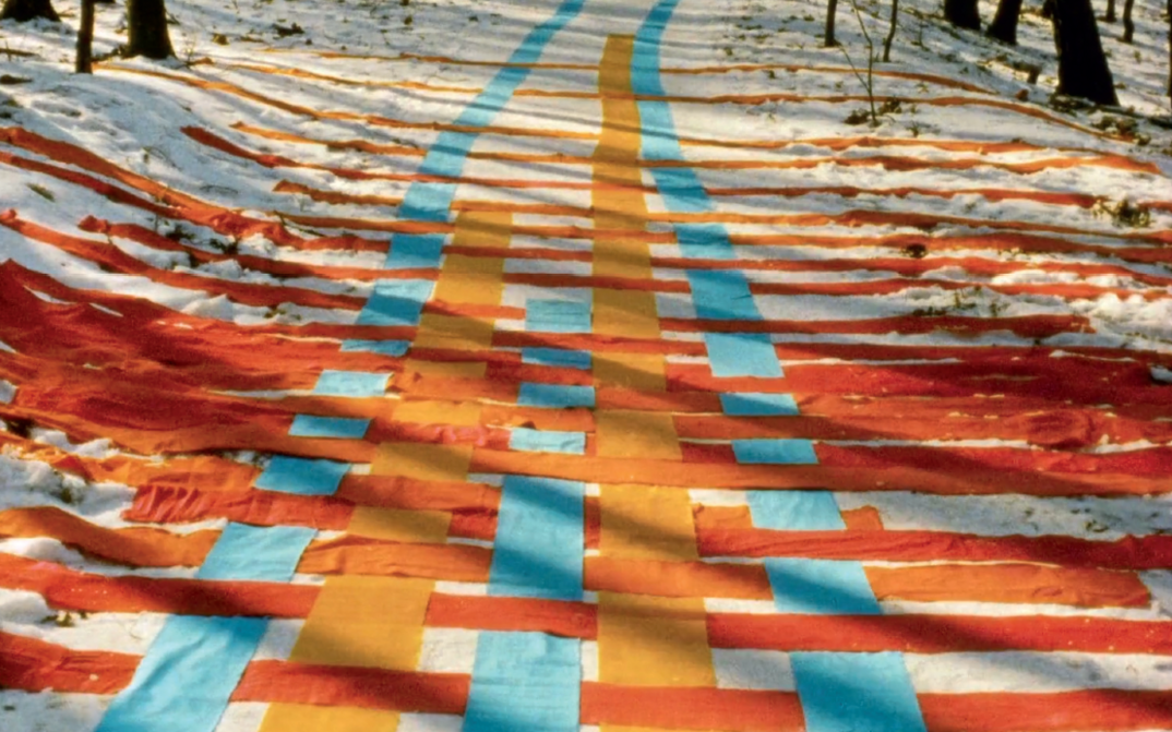Film still from FADENSPIELE: A series of ribbons in yellow, orange and blue are intertwined on a snowy forest path.
