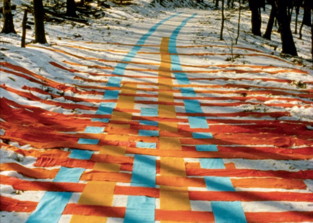 Film still from FADENSPIELE: A series of ribbons in yellow, orange and blue are intertwined on a snowy forest path.
