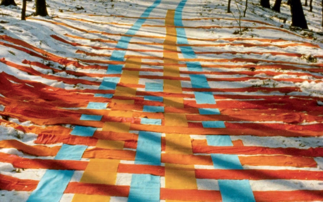 Filmstill aus FADENSPIELE: Eine Reihe von Bändern in Gelb, Orange und Blau sind ineinander verflochten auf einen verschneiten Waldweg gelegt.