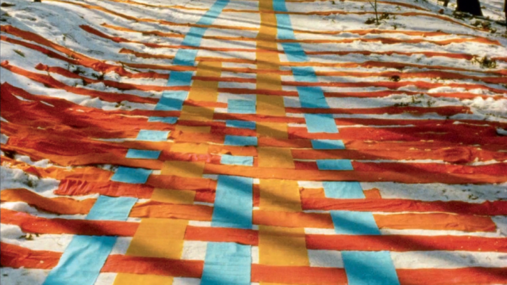 Filmstill aus FADENSPIELE: Eine Reihe von Bändern in Gelb, Orange und Blau sind ineinander verflochten auf einen verschneiten Waldweg gelegt.