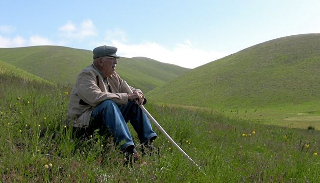 Still from the film "Dilim dönmüyor – Meine Zunge dreht sich nicht" by Serpil Turhan. A man sits among green grassy hills. 