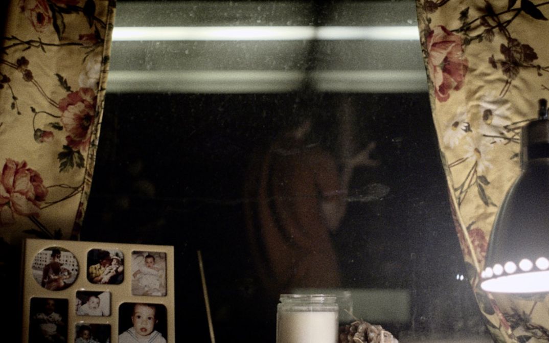 Still from the film "Happer’s Comet" by Tyler Taormina. A window sill at night: family photos in a frame, a candle, a lamp. In the background we see the reflection of a person in an orange shirt. 