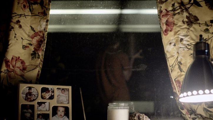 Still from the film "Happer’s Comet" by Tyler Taormina. A window sill at night: family photos in a frame, a candle, a lamp. In the background we see the reflection of a person in an orange shirt. 
