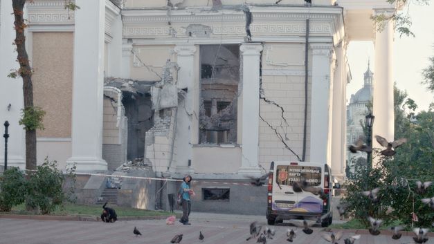 Film still from WHEN LIGHTNING FLASHES OVER THE SEA by Eva Neymann. The image shows a sealed off building with cracks and holes. A person is walking in front of it and pigeons are flying.