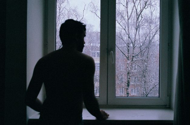A shirtless man in an a slightly out-of-focus silhouette looks out the window at snow falling in front of trees and apartment buildings.